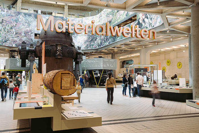 View into the “Material Worlds” exhibition featuring historical industrial objects, a large title sign, and interactive exhibits at the Technical Museum.