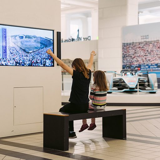 Two children sit on a bench in an exhibition space watching a racing scene on a screen. In the background, a Formula E race car and a grandstand display are visible.
