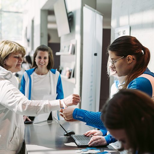 A woman receives a wristband for the familiday at the reception desk from a staff member; in the background, other assistants in blue clothing welcome guests.